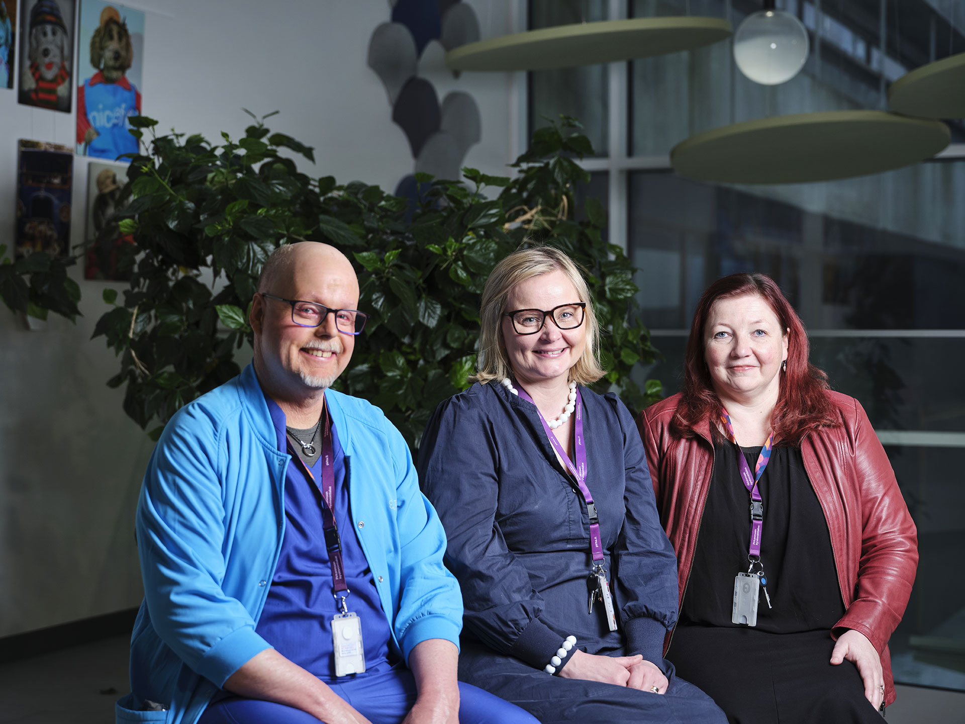Kolme ihmistä istuu vieretysten penkillä Tampereen yliopistollisessa sairaalassa, taustalla viherkasvi.